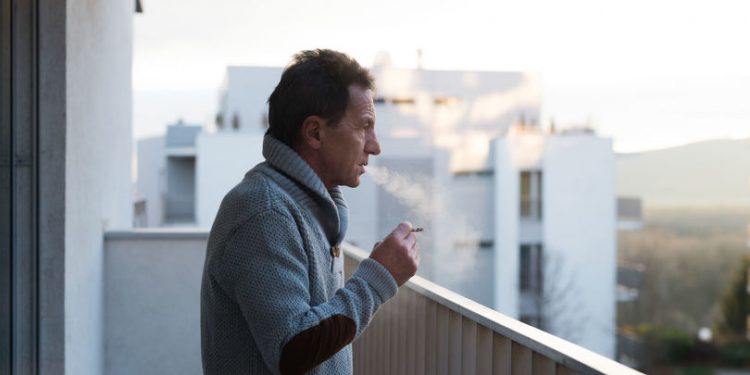Serious senior man standing on balcony and smoking a cigarette