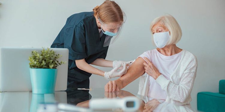 Masked medical nurse holding syringe and doing injection of vaccine to elderly woman at home